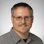 Andrew Gutshall, a man with glasses, short brown hair, and a goatee, wearing a gray plaid shirt, smiles at the camera.