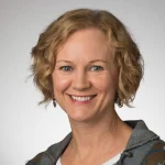 Smiling woman with short curly blonde hair, wearing a gray jacket and silver earrings, on a light background—Christine Paquin.