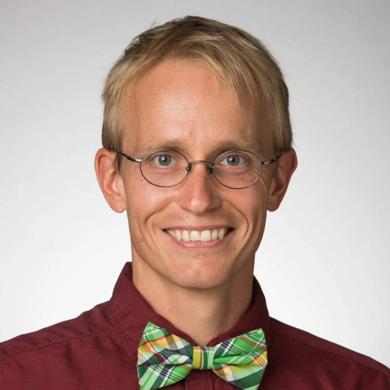 Smiling man with glasses, blond hair, maroon shirt, and a green plaid bow tie, Jon Paul McCool, Ph.D., stands against a light background.