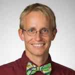 Smiling Jon Paul McCool, Ph.D., with glasses, blond hair, a maroon shirt, and a green plaid bow tie against a gray background.