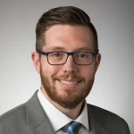 Man with glasses and beard, wearing a gray suit and blue tie, smiling at the camera against a gray background.