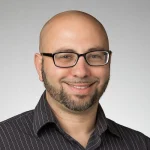 Smiling man with glasses, a beard, and a bald head, wearing a dark striped shirt against a light background.