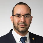 Ossama Abdelgawwad, M.A., a man with short hair, glasses, and beard in a suit and tie, poses confidently against a plain background.