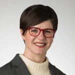 Amanda Brobst-Renaud, Ph.D., with short brown hair, red glasses, a gray blazer, and cream sweater, smiles at the camera.