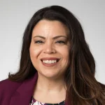 Jessica Bruskoski, a smiling woman with long brown hair, wears a maroon blazer and floral top against a light background.
