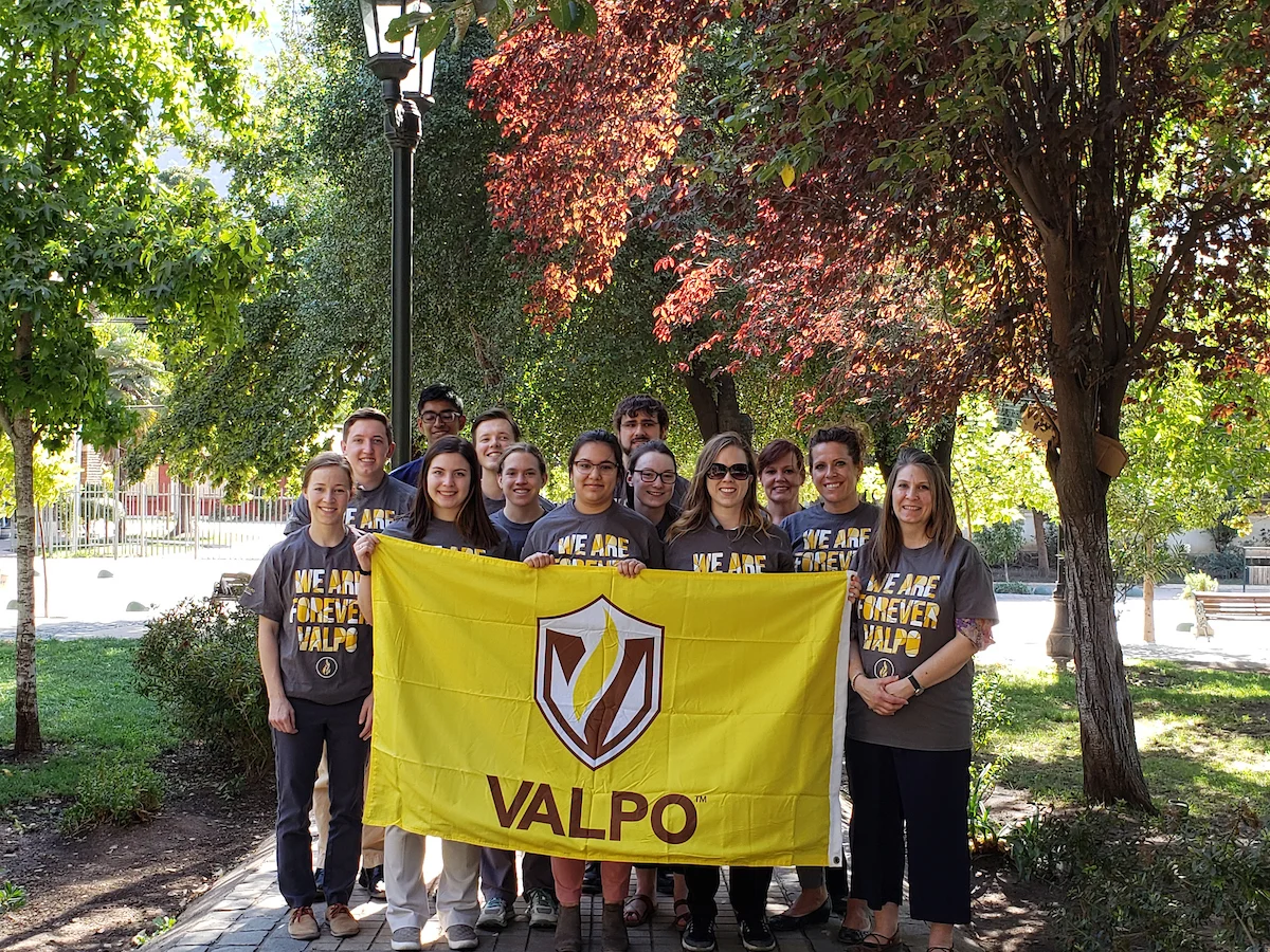 A group of people holds a yellow VALPO flag outdoors, all wearing matching "We Are Forever Valpo" shirts, proudly representing Valparaiso University and abiding by the Valparaiso University Travel Policy.