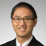 Man with short dark hair and glasses, smiling, wearing a suit and tie against a neutral background.