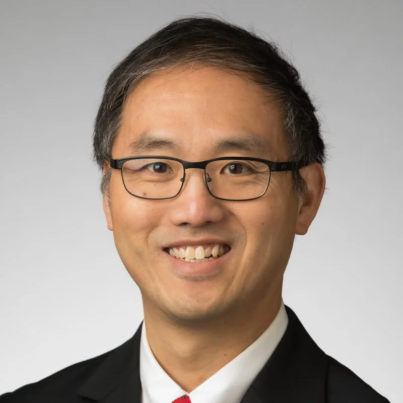 Man with short dark hair and glasses, smiling, wearing a suit and tie against a neutral background.