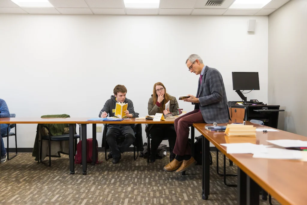 A teacher reads from a book to two students in a classroom, seated at tables with books and notebooks, furthering education opportunities for young learners.