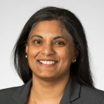 Nirupama Devaraj, a smiling woman with long dark hair, wears a gray blazer while posing confidently in front of a plain light background.