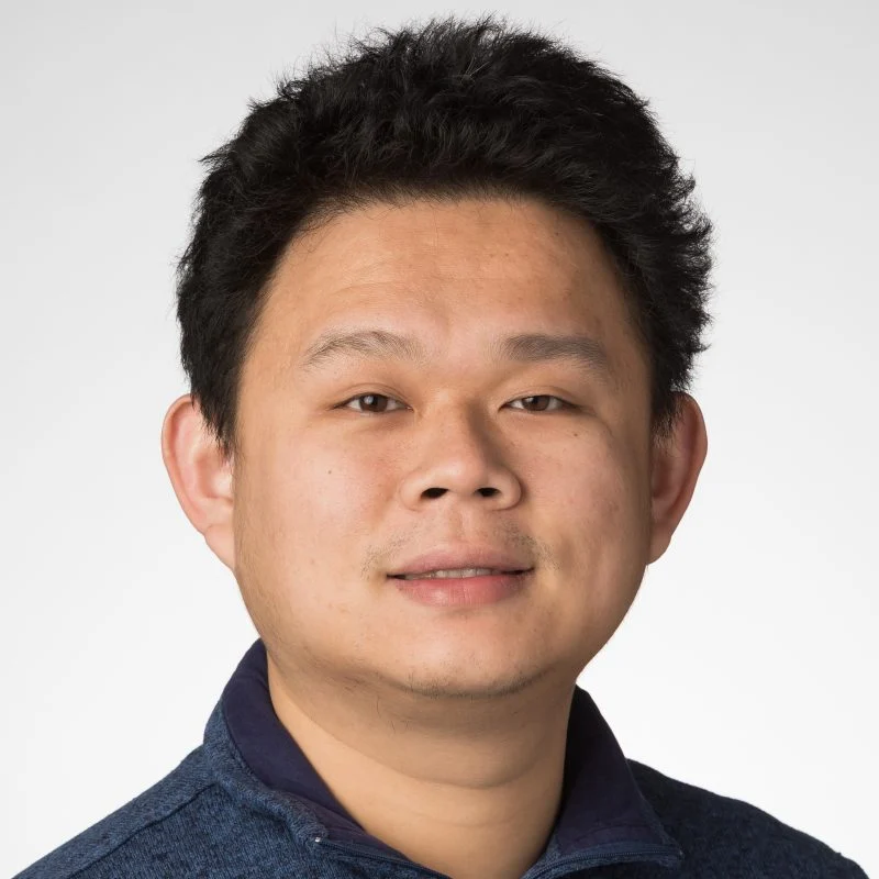 A man with short black hair smiles slightly, wearing a blue collared shirt against a plain background.