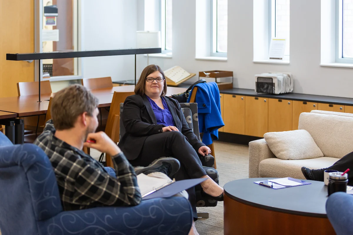 Three people sit and talk in a modern, well-lit office lounge with papers and drinks on the table, discussing academic policies.