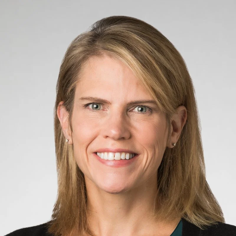 Smiling woman with straight blonde hair, wearing a dark top, in front of a plain light background.
