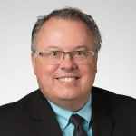 Smiling man in glasses, suit, and blue shirt with a black tie, Dan Maguire, Ph.D., stands in front of a plain light background.
