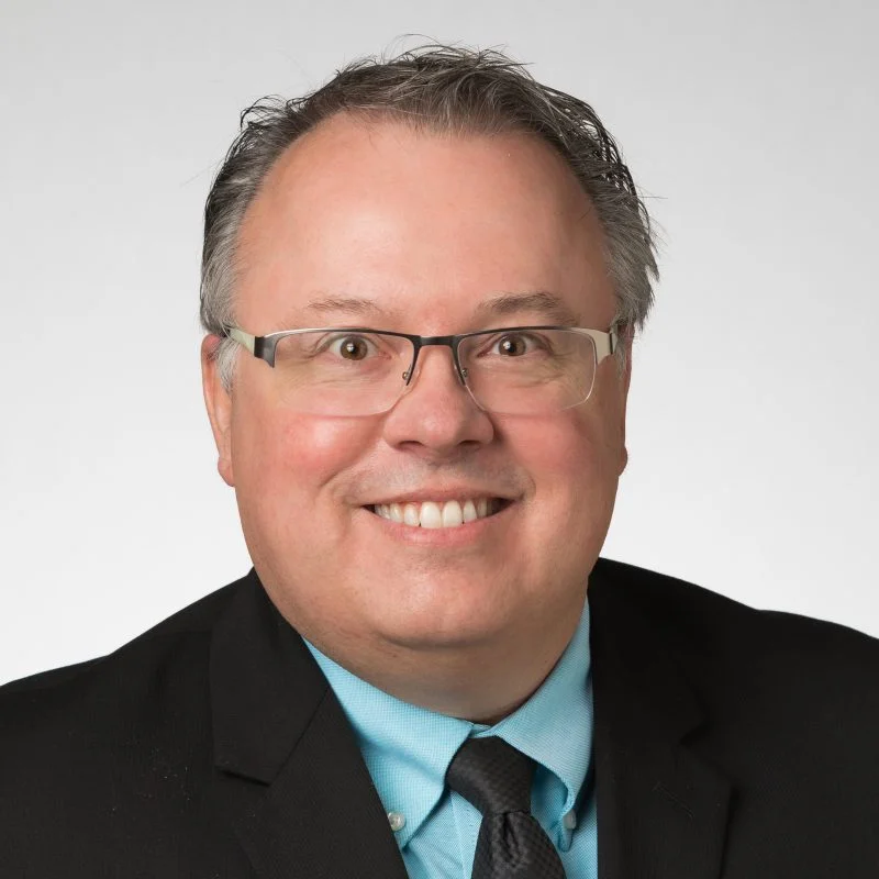 Smiling man in glasses, suit, and blue shirt with a black tie, Dan Maguire, Ph.D., stands in front of a plain light background.
