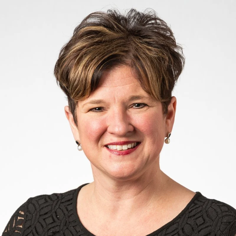 Smiling woman with short brown hair wearing a black top and pearl earrings, Claudine Ruzga poses against a plain background.