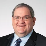 Smiling man with glasses, P.E. and Ph.D., Doug Tougaw, in a suit and tie against a plain light background.