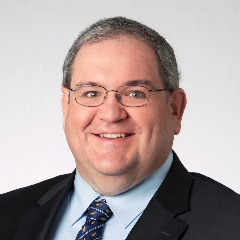 Smiling man with glasses, P.E. and Ph.D., Doug Tougaw, in a suit and tie against a plain light background.