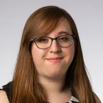 Gwendolyn Kozak, MBA, with long brown hair and glasses, smiles softly in front of a plain light background.