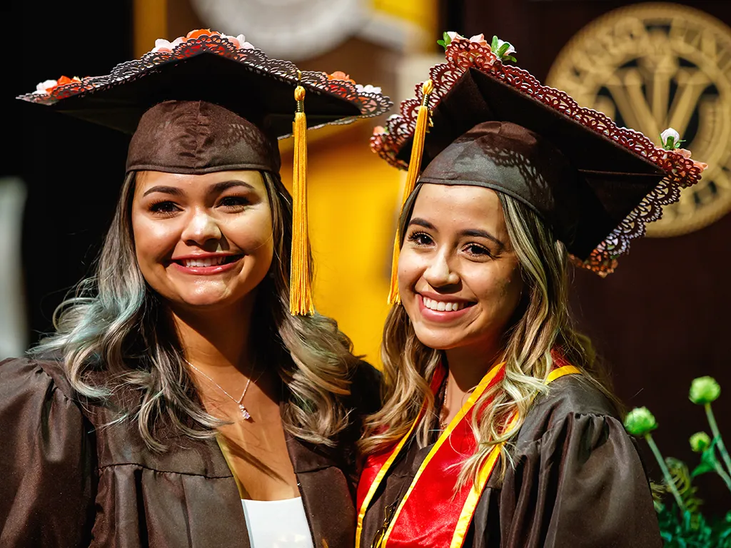 Two graduates in caps and gowns smile at a graduation ceremony, proudly displaying Our Logos on their attire.