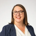 A woman with glasses and shoulder-length brown hair smiles in a blazer and white top against a plain background.