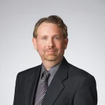 Joseph Bognar, a man with light brown hair and a beard, is pictured wearing a dark suit, gray shirt, and striped tie against a gray background.