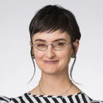 Janneal Gifford, with short dark hair, wearing small glasses, a striped shirt, and jewelry, smiles warmly at the camera.