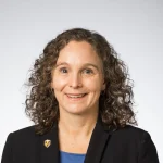 Smiling woman with curly hair wearing a black blazer and blue shirt, posed against a plain background—Karen Hernes.