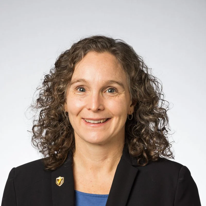 Smiling woman with curly hair wearing a black blazer and blue shirt, posed against a plain background—Karen Hernes.