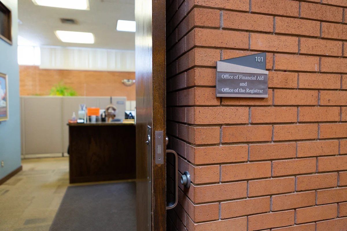 An open door reveals the Office of Financial Aid and Office of the Registrar in a brick-walled building, where students can inquire about grants and other support services.