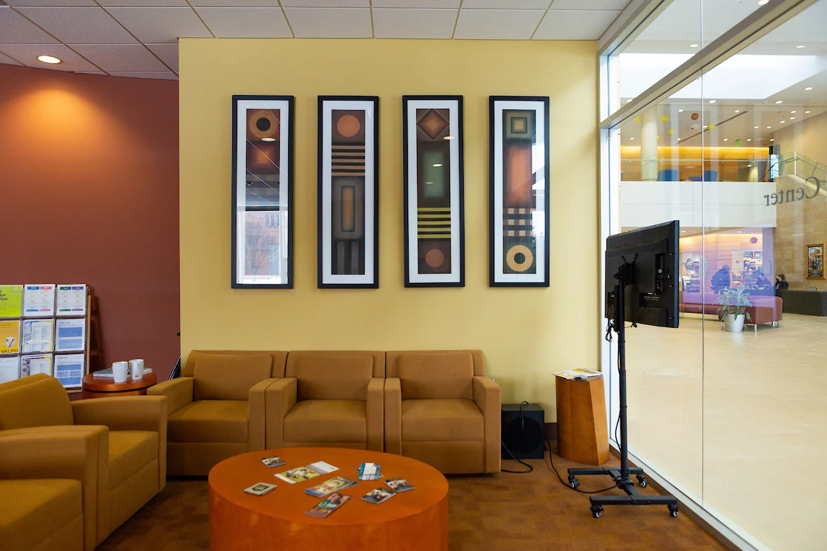 Modern lounge with tan chairs, a round table, and four geometric artworks on a yellow wall near large glass windows, designed to host grants meetings in an inspiring setting.