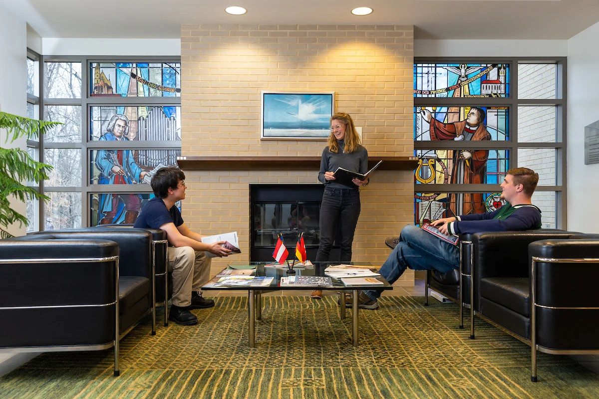 Three people talk in the Kade-Duesenberg German House lounge, with stained glass windows and a fireplace, sitting and standing around a coffee table.