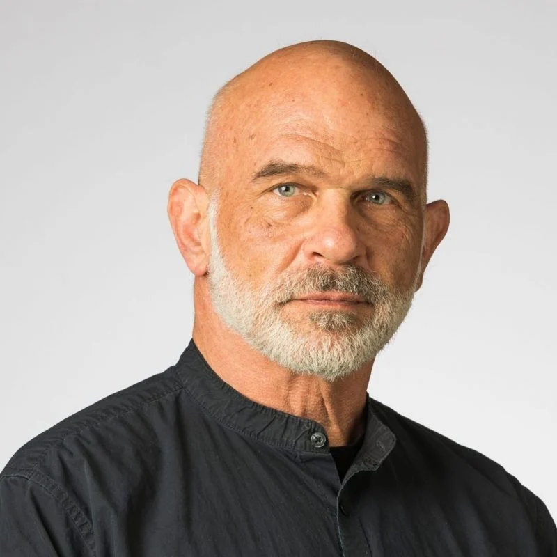 Bald man with a gray beard and black shirt, resembling Shodhin Geiman, Ph.D., looks seriously at the camera against a plain background.