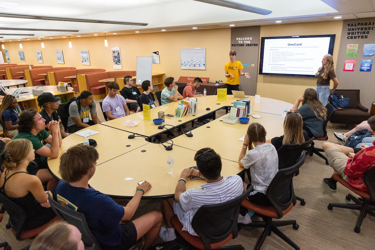 Students sit around a table listening to a presentation in a college classroom, with Student Resources displayed on the screen at the front.