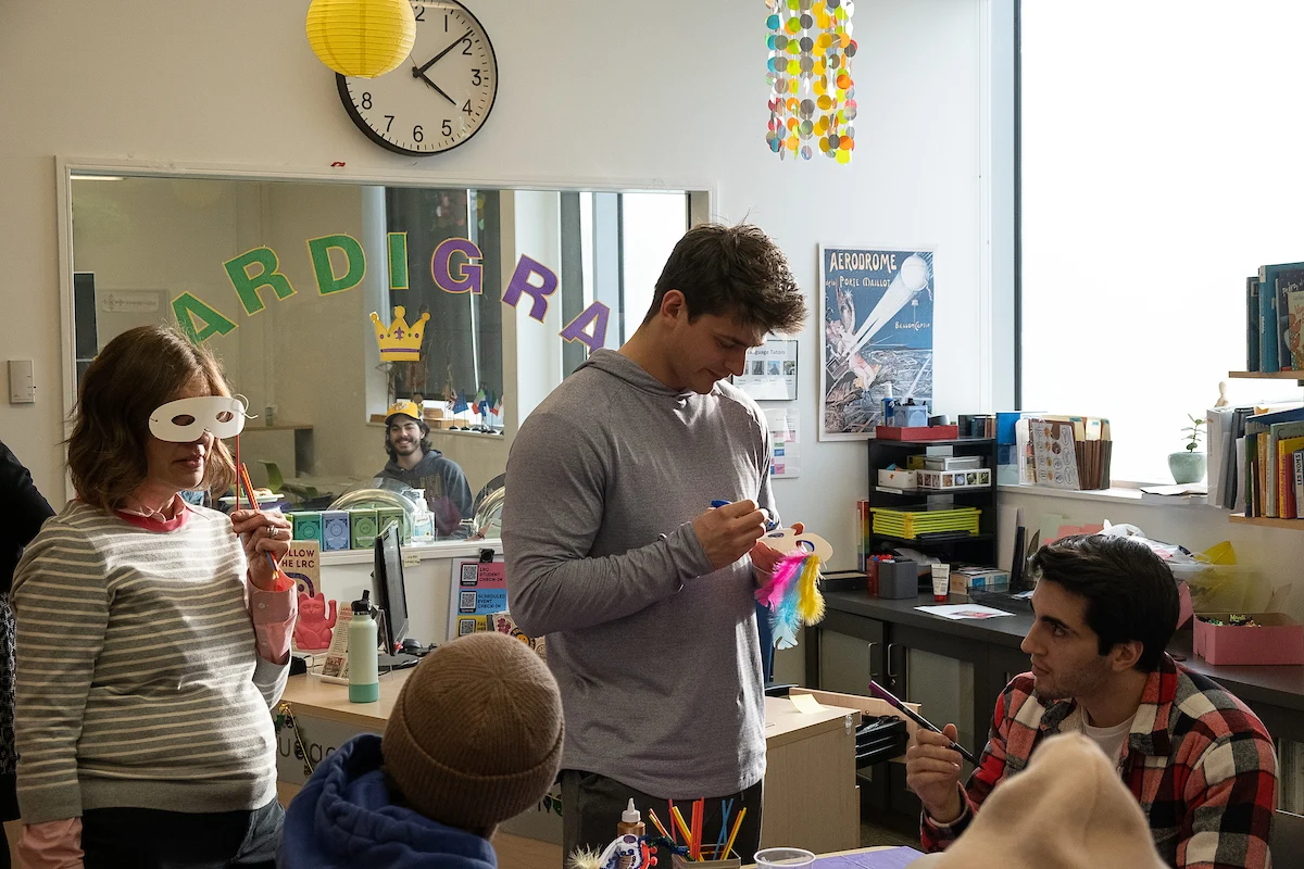 Four adults enjoy making crafts together in a classroom decorated for Mardi Gras, surrounded by colorful art supplies and masks, as part of engaging creative services.