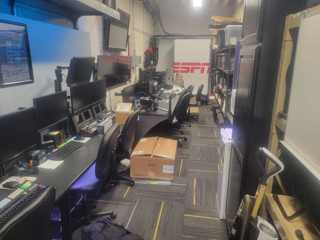 A narrow room with several desks, monitors, and equipment, featuring an ESPN Valparaiso University Productions sign at the far end.