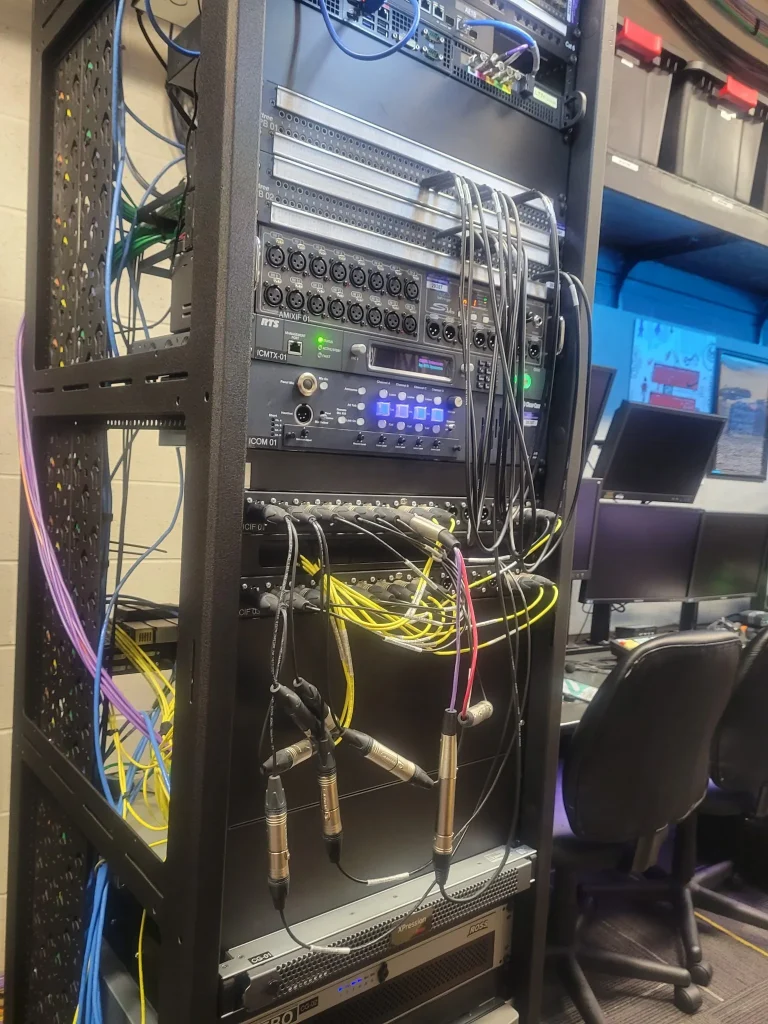 A server rack with connected cables and equipment stands next to desks with monitors in a workspace at ESPN Valparaiso University Productions.