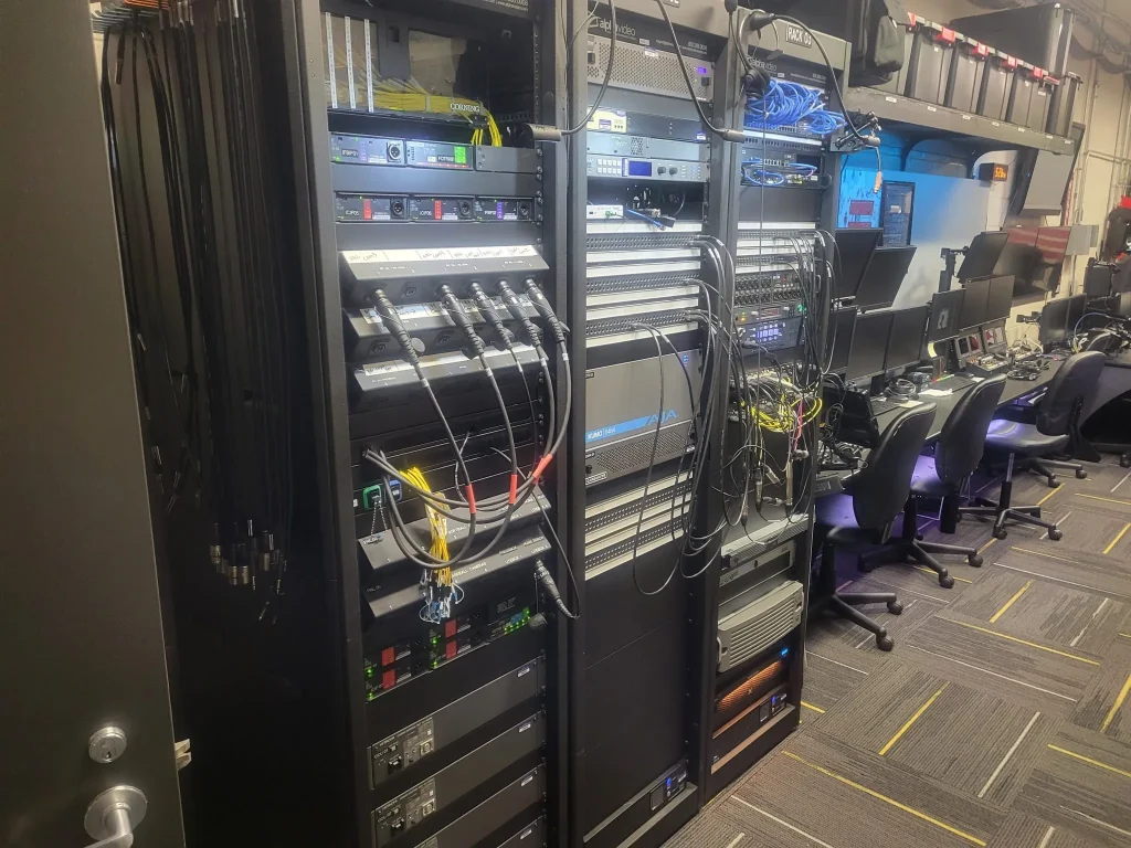 Server room with network racks, cables, and computers; office chairs are lined up along desks on the right, creating a workspace reminiscent of ESPN Valparaiso University Productions.