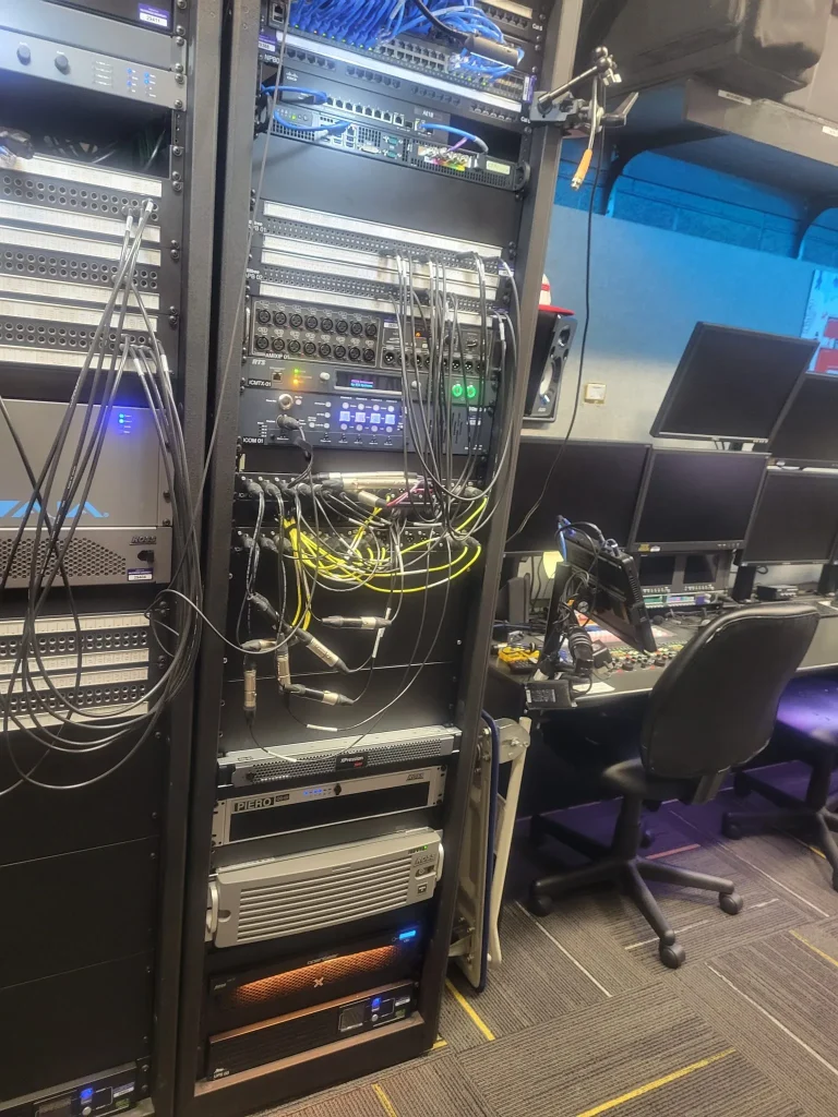 Server racks with tangled cables beside a desk with multiple monitors and office chairs in the ESPN Valparaiso University Productions control room.