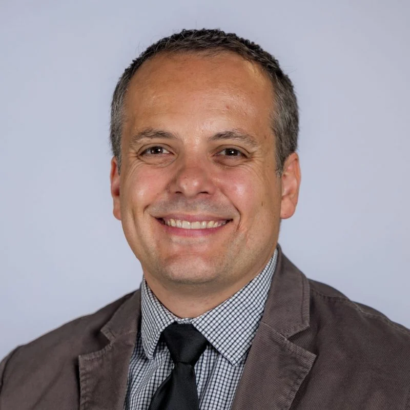 Smiling man, Benjamin Boche, in a gray jacket, checked shirt, and black tie against a plain light background.