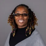Smiling woman with beaded braids, glasses, and a grey shawl, posing in front of a plain grey background—Erica Allen, DNP, RN.