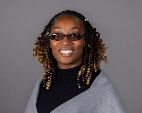 Smiling woman with beaded braids, glasses, and a grey shawl, posing in front of a plain grey background—Erica Allen, DNP, RN.