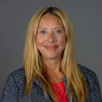 Smiling woman with long blonde hair in a gray blazer and red top, posing against a plain gray background—Jennifer Smolnicky.