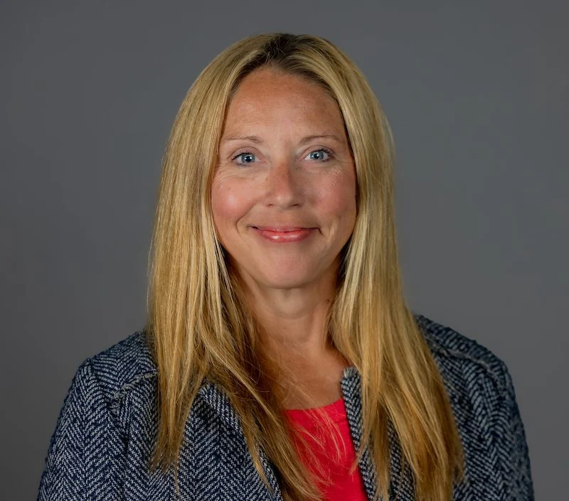 Smiling woman with long blonde hair in a gray blazer and red top, posing against a plain gray background—Jennifer Smolnicky.