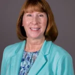 Theresa Kessler, a smiling woman with red hair in a teal blazer and floral blouse, poses against a plain gray background.
