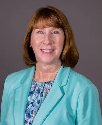 Theresa Kessler, a smiling woman with red hair in a teal blazer and floral blouse, poses against a plain gray background.