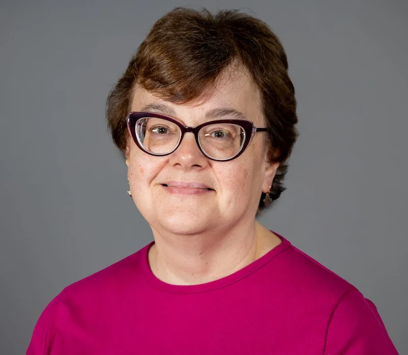 Smiling woman with short brown hair, glasses, and a magenta top against a gray background—Christina Cavinder.