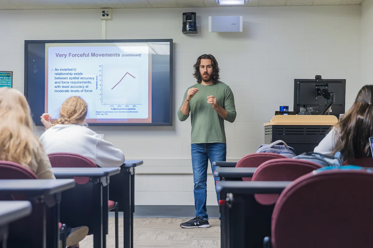 A man teaches a college class about forceful movements using a projected graph and notes, while also referencing relevant academic policies.
