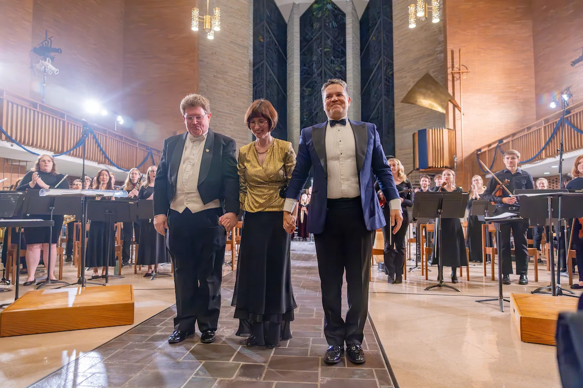 Three formally dressed people hold hands and stand before an orchestra in a concert hall, celebrating achievements shaped by strong academic policies.