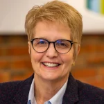 Smiling woman with short blonde hair, glasses, and a blazer, stands in front of a blurred background—Deborah Singer, MBA.